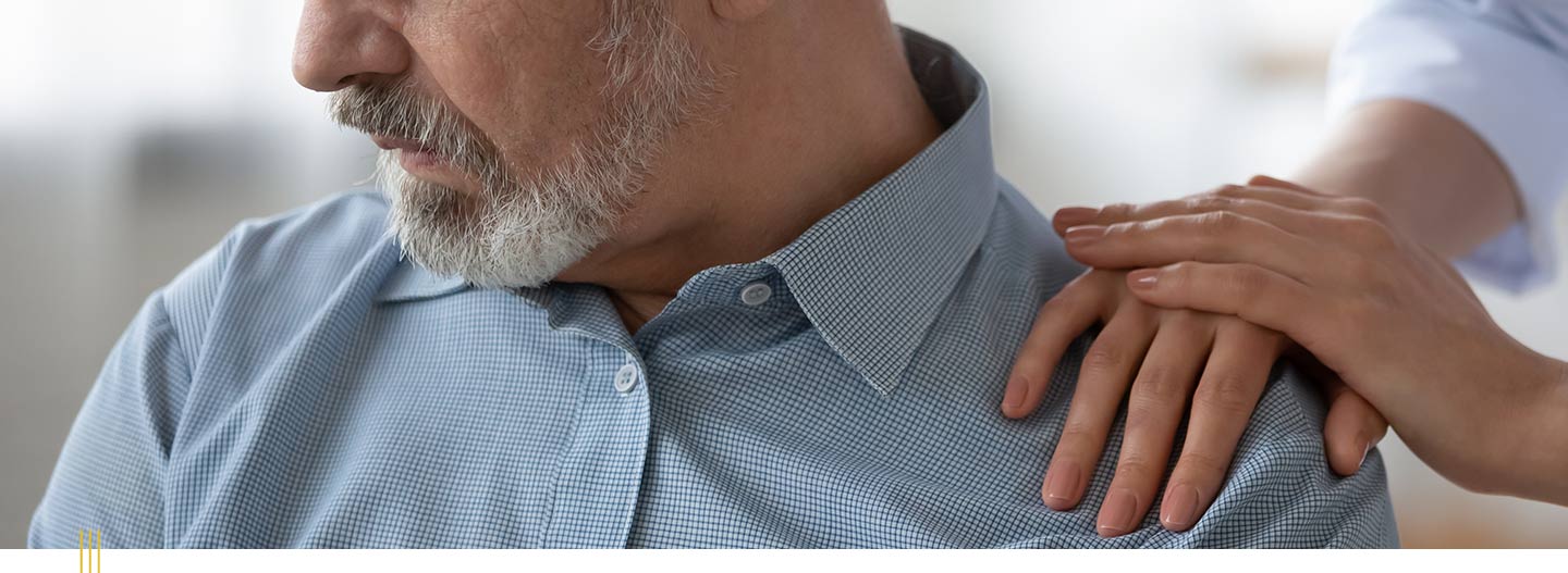 A veteran in a blue checkered shirt looking down while a supportive hand rests on his shoulder.